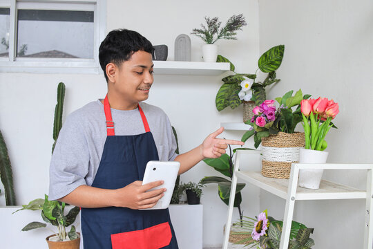 Male Florist In An Apron Holding Tablet Computer While Pointing At Potted Flowers