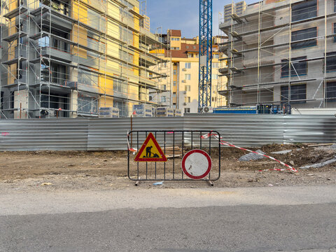 A temporary roadwork barrier with warning and restriction signs stands beside residential buildings under construction with scaffolding and a tower crane.