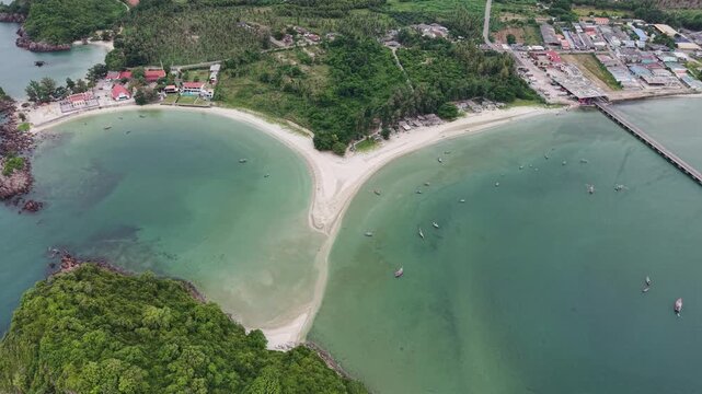 Magnificent aerial view revealing a unique curved sandbar dividing clear tropical waters near a coastal village and long wooden pier in Bang Saphan, Prachuap Khiri Khan, Thailand