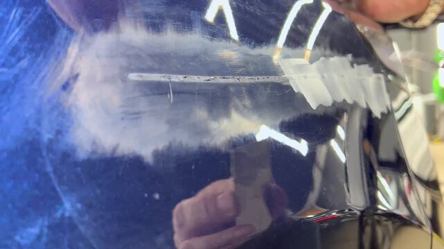 Professional auto mechanic man hands applying polyester car body filler putty with a steel spatula knife tool on a damaged blue vehicle part inside a detailing studio. Professional vehicle bodywork. 