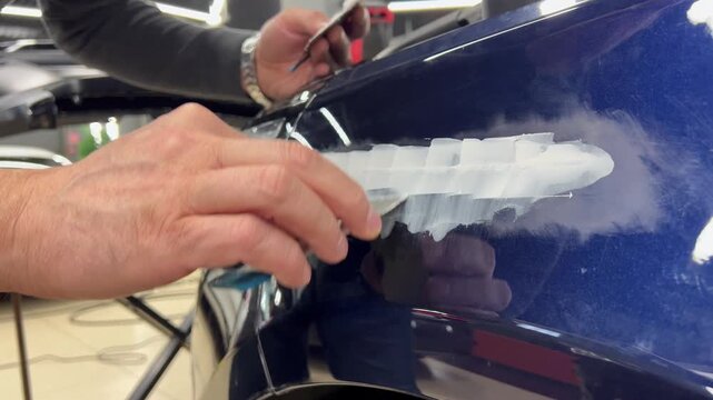 Professional auto mechanic man hands applying polyester car body filler putty with a steel spatula knife tool on a damaged blue vehicle part inside a detailing studio. Professional vehicle bodywork. 