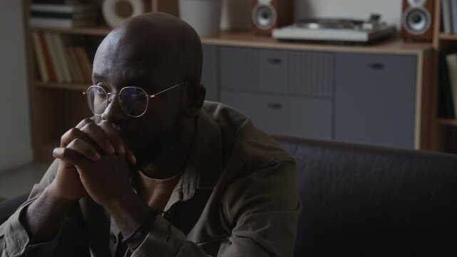 Middle aged African American man in glasses sitting with clasped hands experiencing stress and emotional burnout indoors. Clip ideal for mental health awareness and therapy campaigns