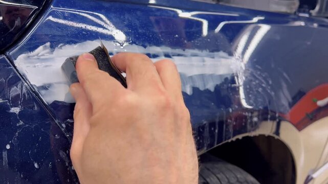 Professional car detailer hands using a sanding block and sandpaper tool for wet sanding body filler on a blue metallic automobile fender. Vehicle bodywork repair, paint preparation, and restoration. 