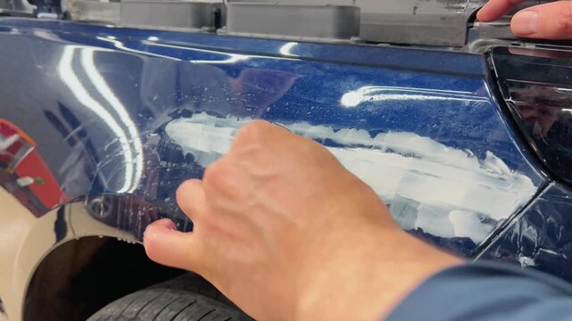 Professional car detailer hands using a sanding block and sandpaper tool for wet sanding body filler on a blue metallic automobile fender. Vehicle bodywork repair, paint preparation, and restoration. 