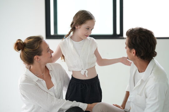 Young parents and their cute child gather on the bench for hugs with their braided haired girl inside the freshly painted empty interior of their future home with copy space