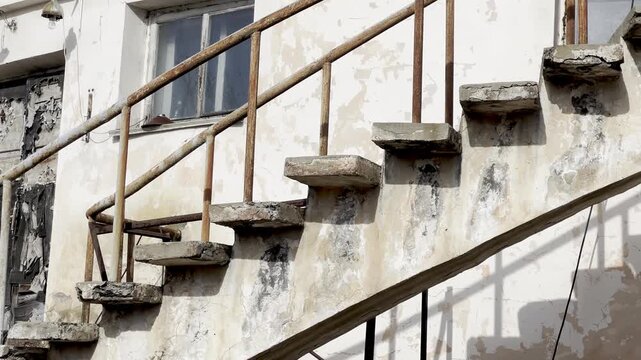 A slow upward pan reveals a weathered concrete staircase with rusted railings ascending a deteriorating white building, highlighting peeling paint, dark stains, and overgrown vegetation at the base.