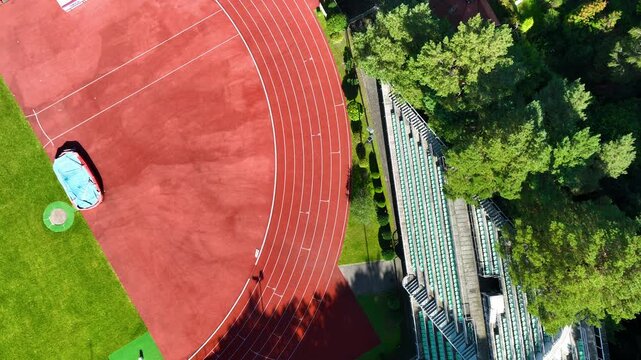 Cinematic aerial topdown shot of a professional red athletics running track and green stadium seating surrounded by lush trees. Perfect for sports, wellness, and fitness content.