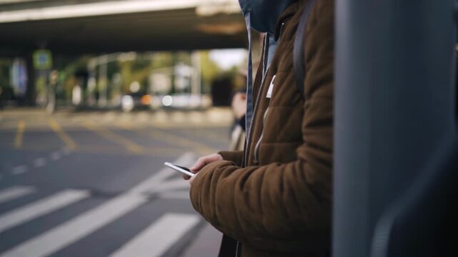 A man in a hoodie and jacket is standing at the intersection and looking at his mobile phone. His face is covered. The shot is taken from the side.