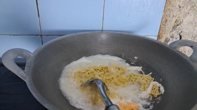 Video footage of cooking instant noodles with soup and ingredients inside a metal wok frying pan on a stove, representing home culinary, quick meal preparation, Asian street food, and hot noodle recip