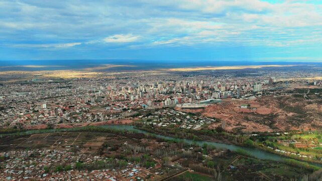 Neuquen city skyline, Limay River, dense residential districts, cultivated valley, eroded barda terrain and Patagonian plateau, drone lateral slide