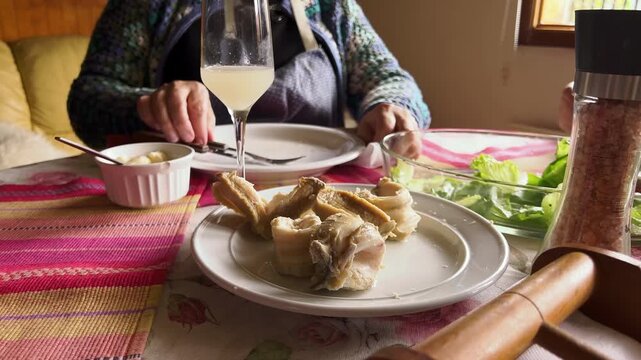 Hands reach across a colorful woven table runner set with tender Chilean loco shellfish, a glass of pisco sour, pink salt, and crisp lettuce. A warm, homemade coastal feast.