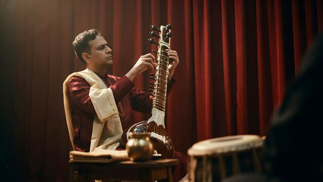 Classical Indian musician playing sitar on stage with traditional instruments and red curtains backdrop