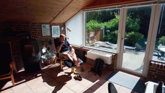 Static wide shot of a man relaxing with coffee in a sunroom armchair overlooking a sunny garden terrace, Scandinavia, Denmark