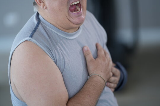Obese senior man with sudden chest pain kneels clutching his heart beside the treadmill after physical exertion in the gym, candid real moment. Cardiovascular health and obesity risk concept