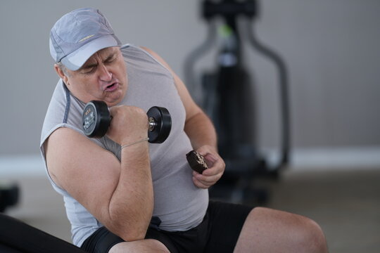Heavyset mature man fighting food temptation munches a donut mid workout, dumbbell still gripped during his weight loss workout at the fitness club, candid real moment