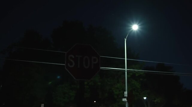 Dark STOP sign beneath bright streetlight on quiet street at night appears among trees and power lines, creating an eerie atmosphere on an empty urban street