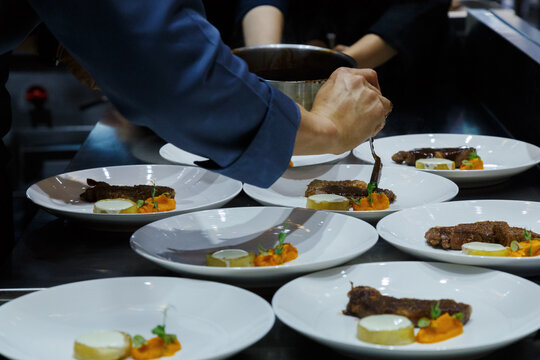 professional chef pouring brown sauce over stack meat decoration dishes during dinner food plating service in commercial restaurant kitchen. food service, hotel and restaurant cooking.