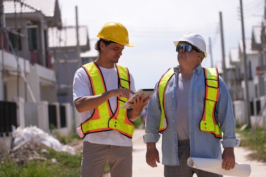 Construction supervision team in helmets and vests walk the site with coffee, pointing out finished units on an inspection walk at a large housing construction project on a bright sunny day
