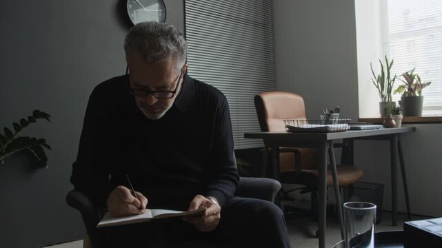 Middle aged Caucasian psychotherapist writing notes in notebook while listening to patient during therapy session. Ideal for mental health awareness and counseling campaigns