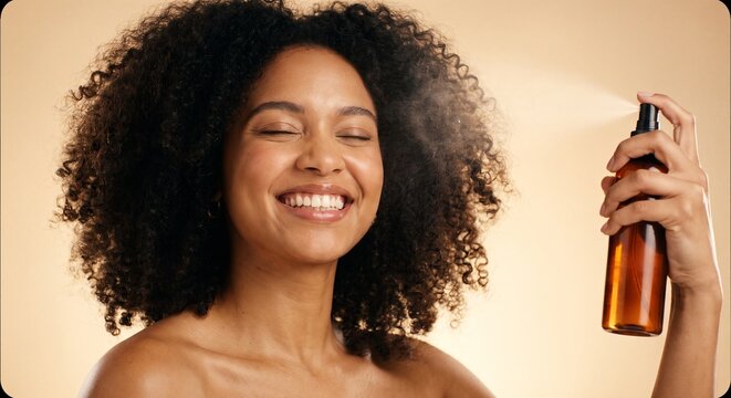 Smiling young woman with curly hair spraying refreshing facial toner from a bottle during her daily beauty skincare routine