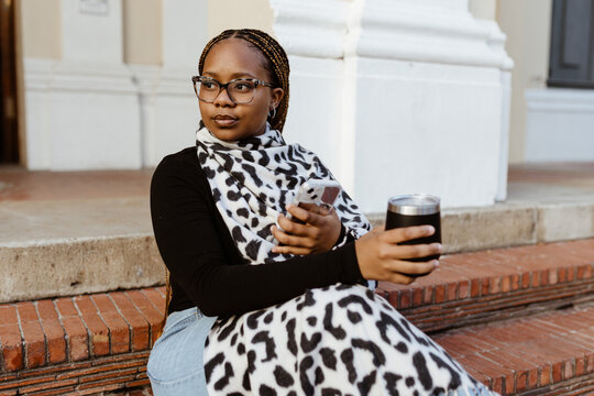 Stylish Black woman with phone and tumbler on city steps