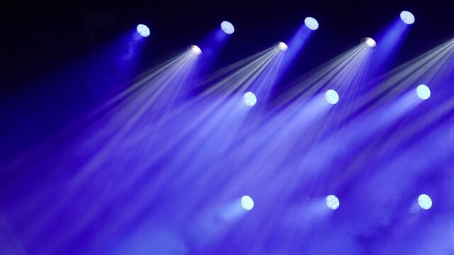 Multiple bright blue and violet spotlight beams shining diagonally in parallel across a dark stage filled with dense fog during a music concert