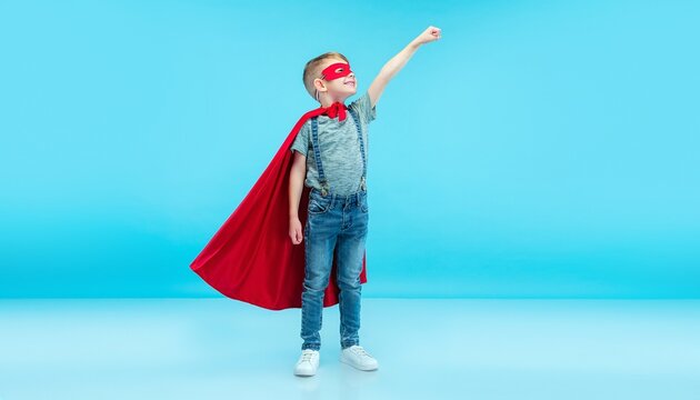 Happy little boy wearing superhero red cape and mask smiling while posing with raised fist up in the air pretending to fly during creative imaginative play