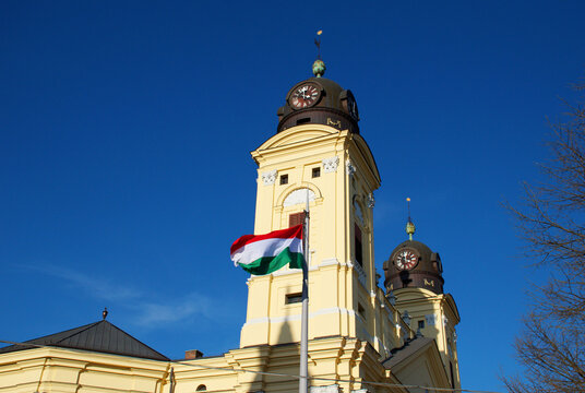 Historic Reformed church in Debrecen, Hungary, photographed against a clear blue sky with the Hungarian national flag displayed on the facade. The ornate yellow Baroque architecture, twin clock towers