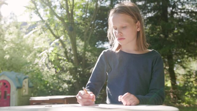 Young student sketches carefully on paper during creative outdoor lesson. Calm girl practices drawing with pencil at garden desk, surrounded by natural greenery under gentle afternoon light.