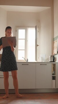 Young woman dancing barefoot in her bright kitchen while holding a laptop, enjoying a happy moment of remote work, representing a modern and flexible lifestyle concept. Vertical video