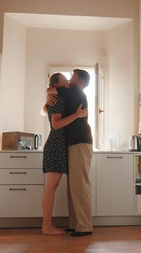 Romantic young couple hugging and kissing while dancing together in a bright, sunlit kitchen, showing their affectionate relationship and shared intimate moment at home. Vertical video