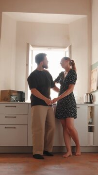 Romantic young couple holding hands and talking while standing together in a sunlit kitchen, enjoying a tender moment of love, intimacy, and connection at home. Vertical video