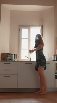 Barefoot young woman in a dress opening a window and drinking coffee in her minimalist kitchen, enjoying a tranquil morning and the bright sunlight. Vertical video