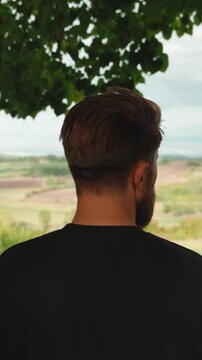 Young bearded man with a stylish haircut admiring the beautiful tranquil countryside landscape from a viewpoint, contemplating and thinking about the future. Vertical video 