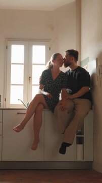 Romantic young couple talking, laughing, hugging and kissing while sitting on the kitchen counter, showing affection and enjoying quality time together in their apartment. Vertical video 