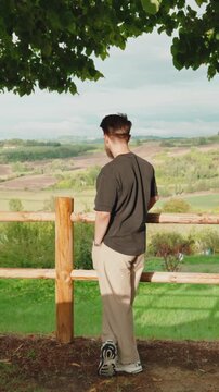 Young man standing near a wooden fence and enjoying the view of the rolling hills of tuscany while the wind blows through his hair in a peaceful natural environment. Vertical video 