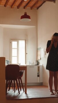 Young woman having an emotional and stressful conversation on her smartphone while pacing anxiously back and forth inside her cozy and sunlit apartment kitchen. Vertical video 