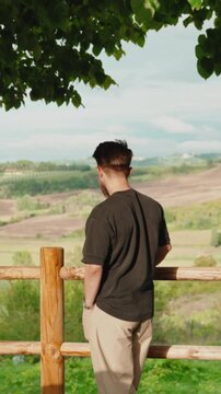 Young man enjoying a panoramic view of the rolling hills and green countryside, contemplating life from a peaceful observation deck in a serene and natural setting. Vertical video 