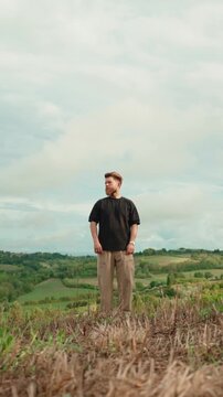 Young handsome man with a beard standing alone in nature on a hilltop, enjoying the peaceful scenic view of the lush green rolling hills and cloudy sky in the background. Vertical video 