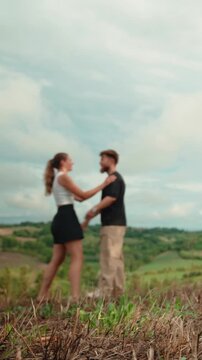 Romantic young couple running, embracing, and kissing on a grassy hill, sharing an intimate moment outdoors with a beautiful scenic landscape of rolling hills and sky. Vertical video 