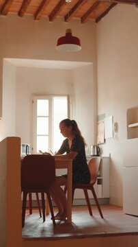 Young woman walking and sitting down at her kitchen table while looking at her smartphone, illuminated by soft natural light coming from the window on a summer morning. Vertical video 