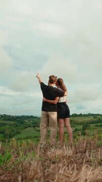 Back view of a young romantic couple hugging and enjoying the scenic view from a hilltop, with the man pointing at the beautiful green landscape stretching out before them. Vertical video 