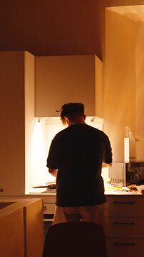 Back view of a solitary man preparing food at night in his modern kitchen, chopping ingredients on a cutting board under a warm light, embracing a cozy and independent domestic lifestyle. Vertical