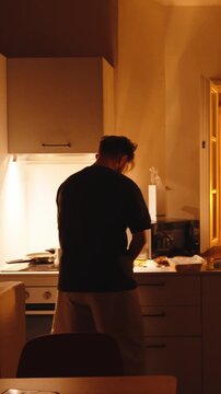 Silhouette of a man from behind dancing and moving rhythmically while cooking a meal in a modern kitchen at home during the evening, creating a cozy and warm atmosphere. Vertical video 
