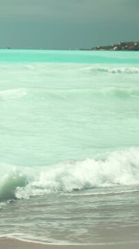 Serene turquoise ocean waves washing over a sandy beach with a distant coastline under a tranquil sky, creating a peaceful and calming atmosphere for relaxation. Vertical video 