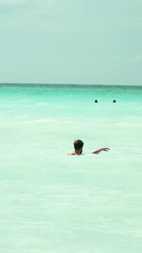 Back view of a shirtless man with a beard swimming and relaxing in the turquoise ocean, enjoying a tropical vacation on a sunny summer day and raising arms in joy. Vertical video 