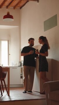 Romantic young couple enjoying morning coffee, talking and laughing together in their cozy sunlit kitchen, sharing an intimate and tender moment with a kiss and smiles. Vertical video 