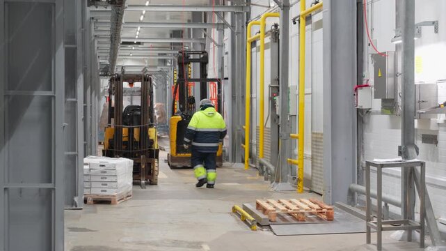 Worker in high-visibility clothing walking through a large industrial warehouse with a forklift