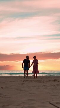 Romantic couple holding hands and kissing while walking on a sandy beach during a beautiful and colorful sunset, enjoying their peaceful honeymoon and vacation together. Vertical video