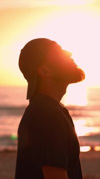 Silhouette of a pensive bearded man raising his head to the sunset on the beach, embodying reflection, hope, and freedom while enjoying a peaceful moment by the sea. Christian man praying. Vertical 